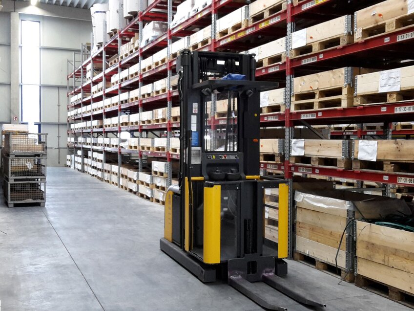 Hochhubstapler in einem Schmalganglager mit hohen Regalen und gestapelten Holzkisten.