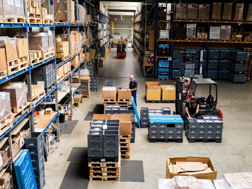 Kommissionierbereich mit hohen Regalen, Mitarbeitern beim Verpacken und einem Gabelstapler zwischen Paletten und Kisten.